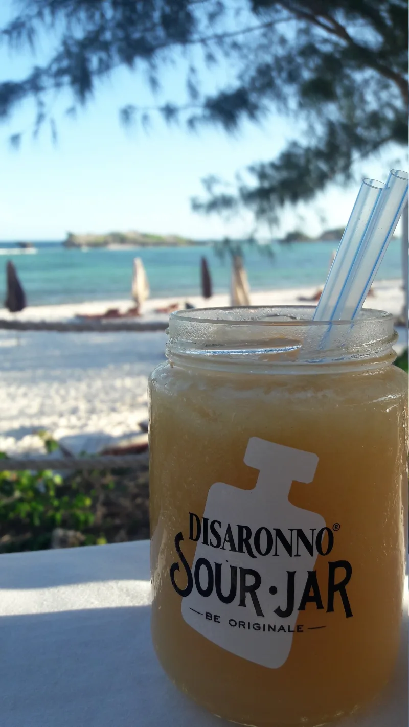 A frosted jar of pale cocktail with two straws on a white tablecloth, with the beach umbrellas and the lagoon behind.