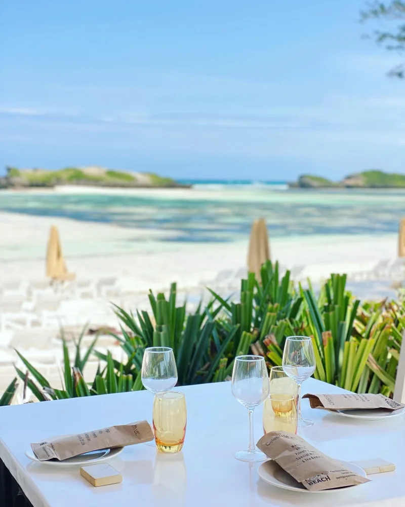 A white table laid with wine glasses and Papa Remo Beach napkins, looking out over closed umbrellas, the sandbar and the lagoon.