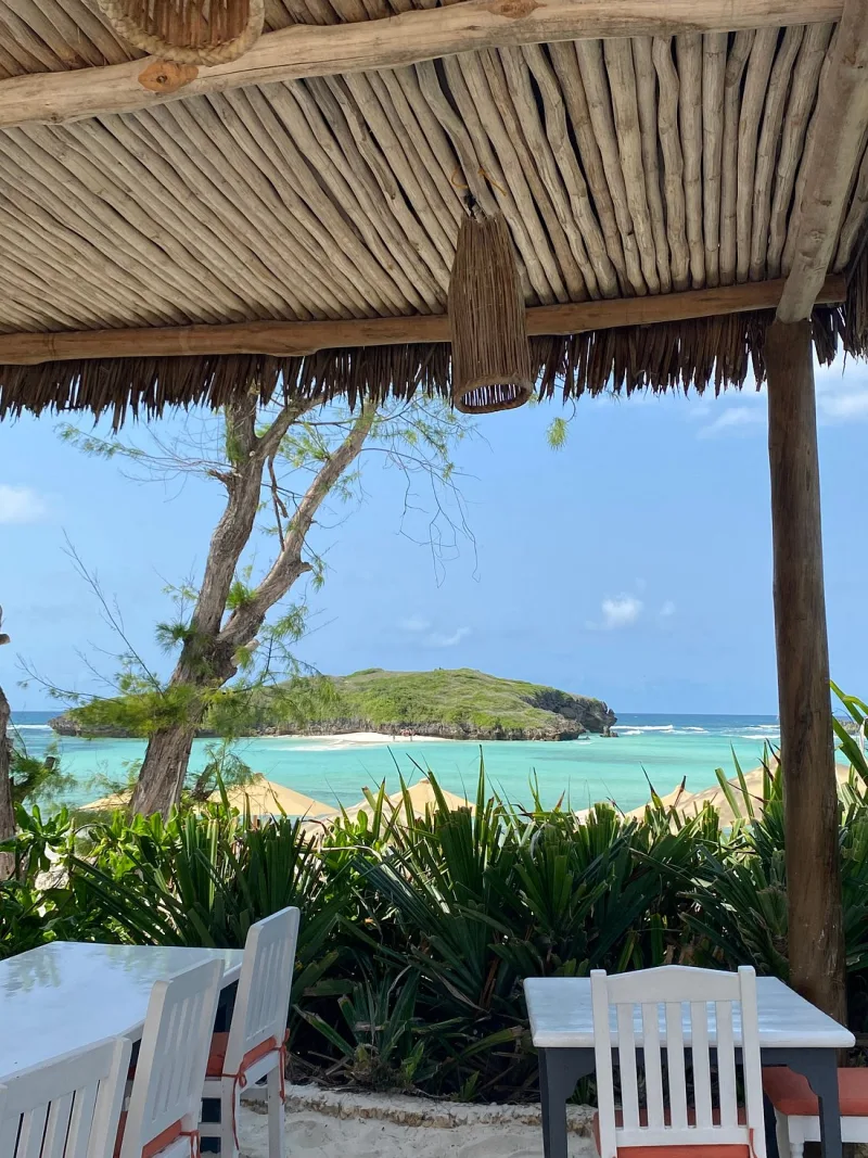 The lagoon and a green islet framed by the edge of a makuti roof and a casuarina tree, with cream umbrellas on the sand below.