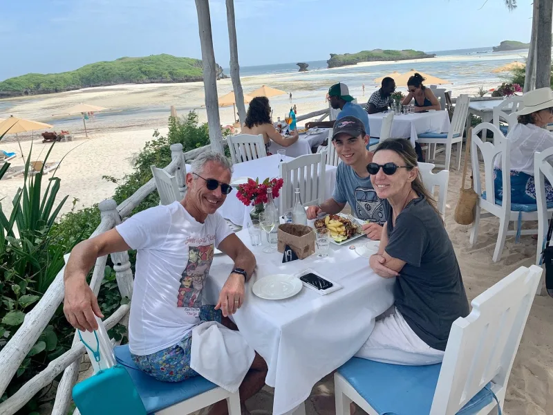 Three guests smiling at a table laid with white linen beside the beach rail, with umbrellas, the lagoon and the islets behind.