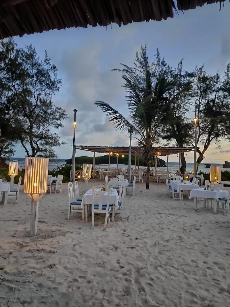 Tables laid for dinner on the sand at dusk, lit by tall white lanterns, with a palm and the sea behind.
