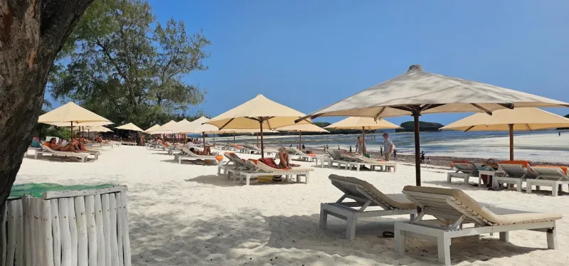 Rows of cream umbrellas and white loungers along the beach with guests sunbathing, and people walking at the water line.