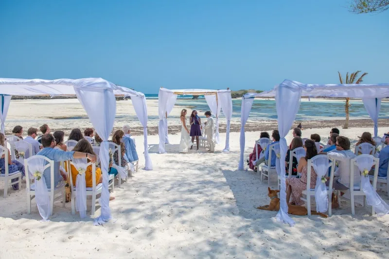 A couple standing with a celebrant under a white draped canopy on the sand, with guests seated on white chairs and the lagoon behind.