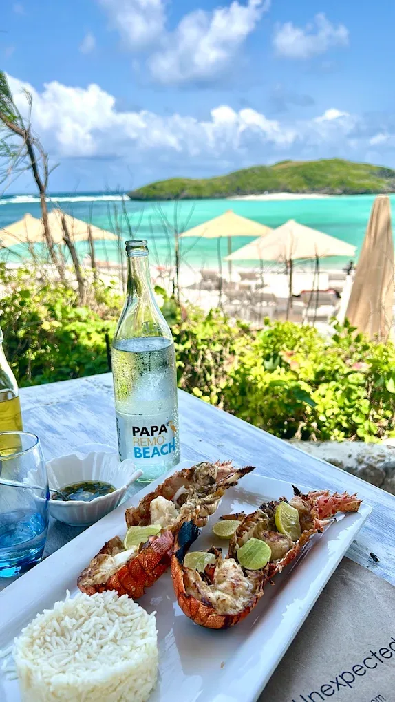 Two halves of grilled lobster with lime and a mound of rice on a white plate, with a Papa Remo Beach water bottle and the lagoon behind.