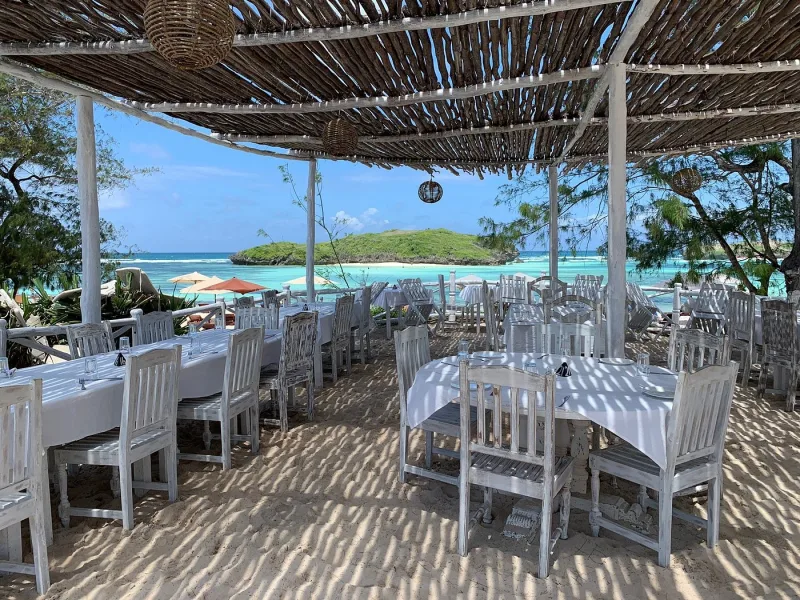 Whitewashed tables and chairs set on the sand under a driftwood and makuti roof, looking out to the lagoon and a green islet.