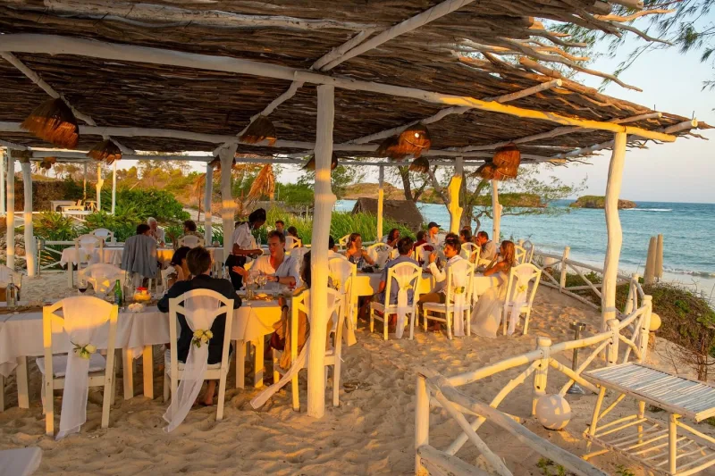 Guests seated at long white tables under a driftwood pergola in low golden light, with the sea beyond.