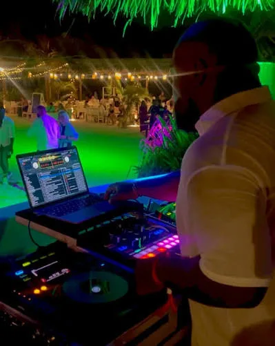A DJ working at a laptop and mixer under coloured lights at night, with guests and festoon lights behind.