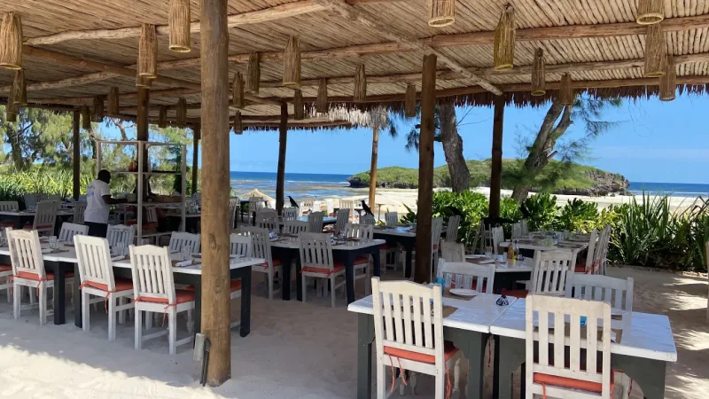 Rows of white tables and chairs with orange cushions under a makuti roof hung with woven lanterns, opening onto the beach.