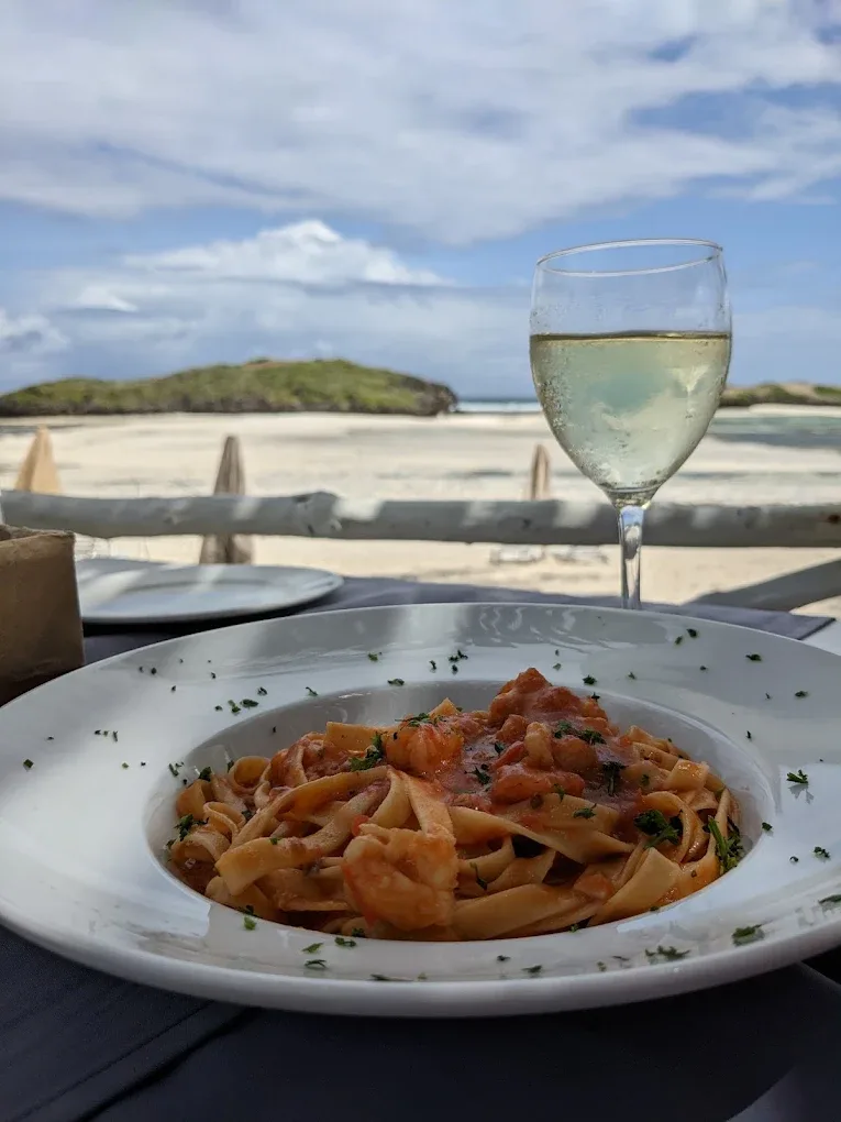 A bowl of tagliatelle with prawns in tomato sauce beside a glass of white wine, on a table looking out to the sandbar.
