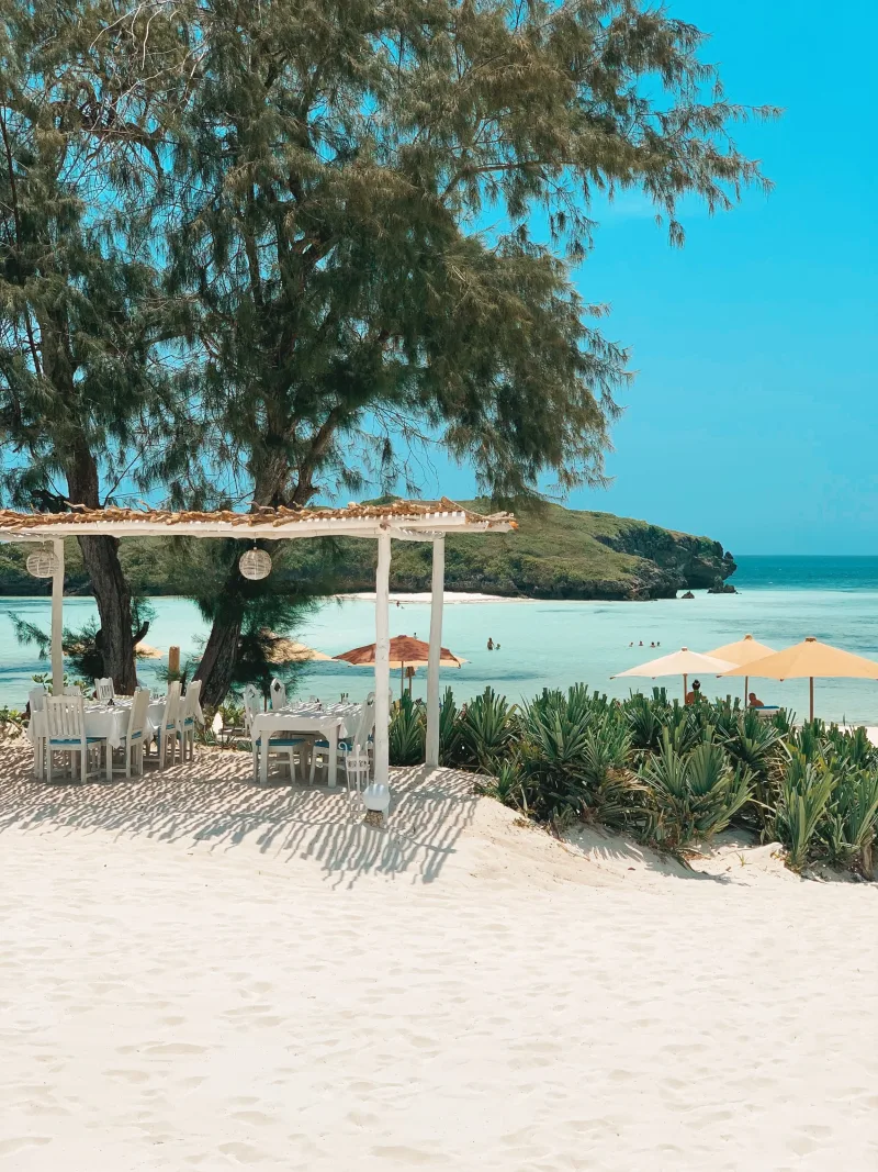A white pergola hung with lanterns over laid tables on the sand, with the lagoon, swimmers and a green headland beyond.