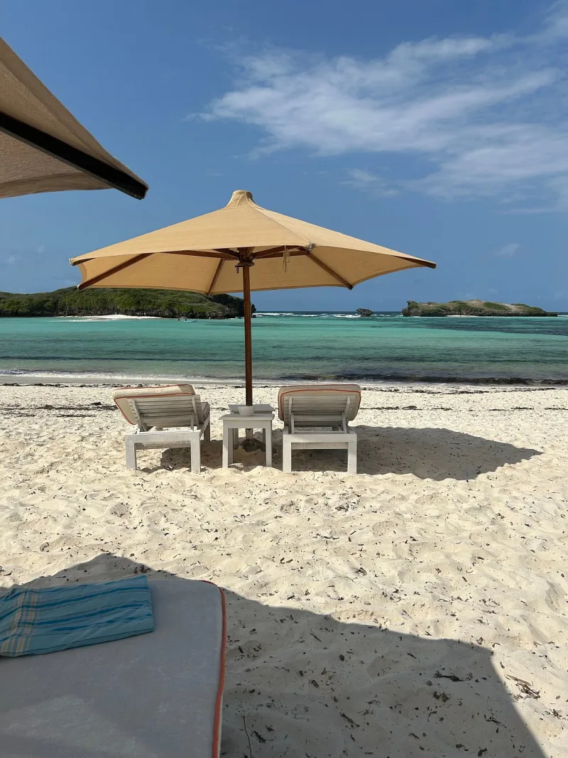Two white sun loungers and a small table under a cream umbrella on the sand, facing the lagoon and the islets.