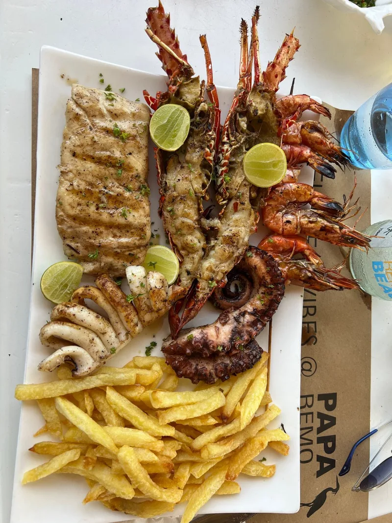 A long white plate of grilled seafood — half lobsters, whole prawns, calamari rings, an octopus tentacle and a fish fillet — with lime wedges and chips.