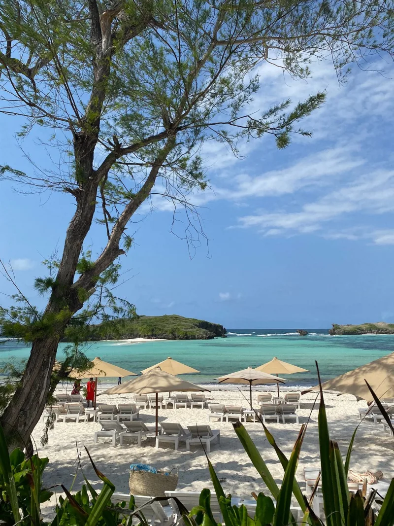 Sun loungers and cream umbrellas on white sand, seen through casuarina branches, with the turquoise lagoon and two green islets beyond and a Maasai askari in a red shuka standing on the sand.