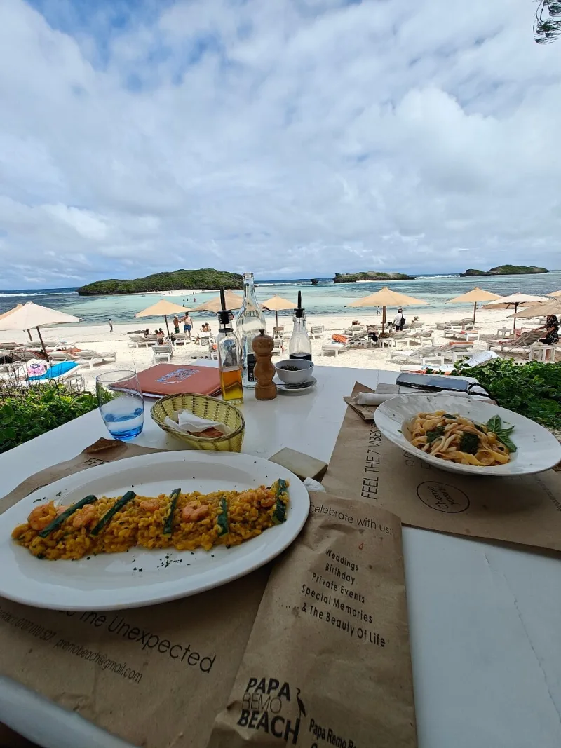 A plate of yellow rice with prawns and a bowl of tagliatelle on a table, looking down over umbrellas, loungers and the lagoon.