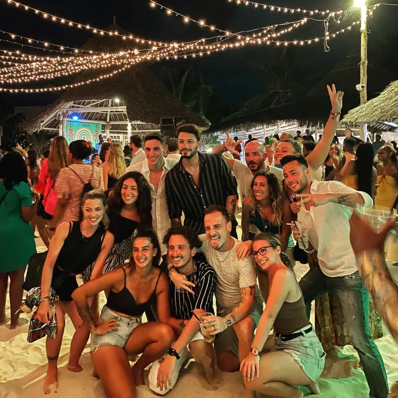 A crowd of guests posing and smiling on the sand at night under strings of festoon lights, with makuti roofs and a lit stage behind.