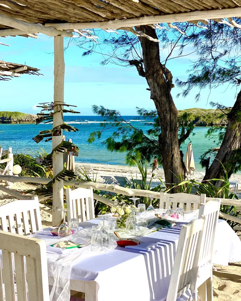 A long table laid with white linen, glasses and flowers under a driftwood roof, with the beach and the reef beyond.