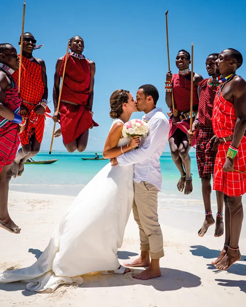 A bride holding a bouquet kissing a groom on white sand, surrounded by Maasai men in red shukas leaping with sticks raised, with turquoise water behind.