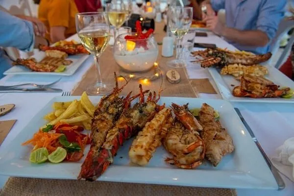 A platter of grilled lobster and prawns with chips and salad on a candlelit table, with a glass of white wine and other diners behind.