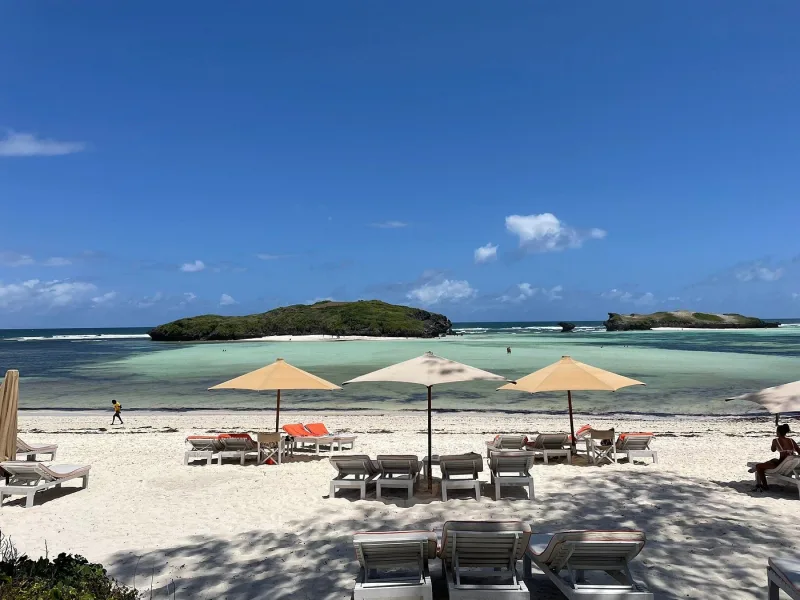 Three cream umbrellas and rows of white sun loungers on the beach, with the shallow turquoise lagoon and two green islets under a bright blue sky.