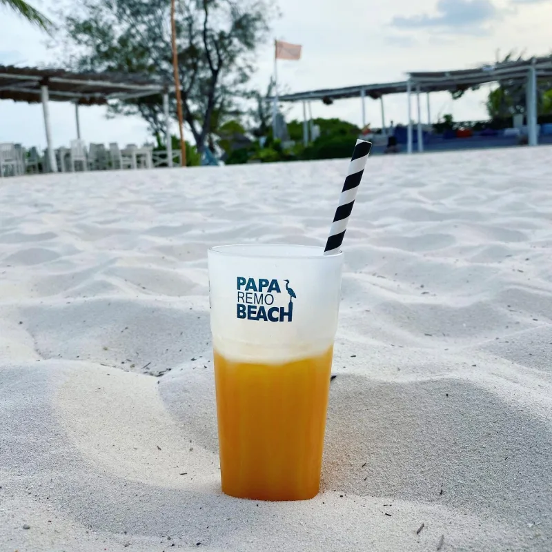 A frosted Papa Remo Beach glass of orange juice with a striped paper straw, standing in the white sand with the beach bar behind.